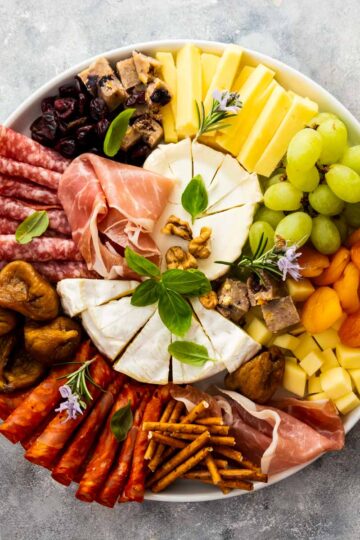 a closeup, overhead shot of a charcuterie board with brie, cubed cheeses, crostini, jam, grapes, dried apricots, and nuts
