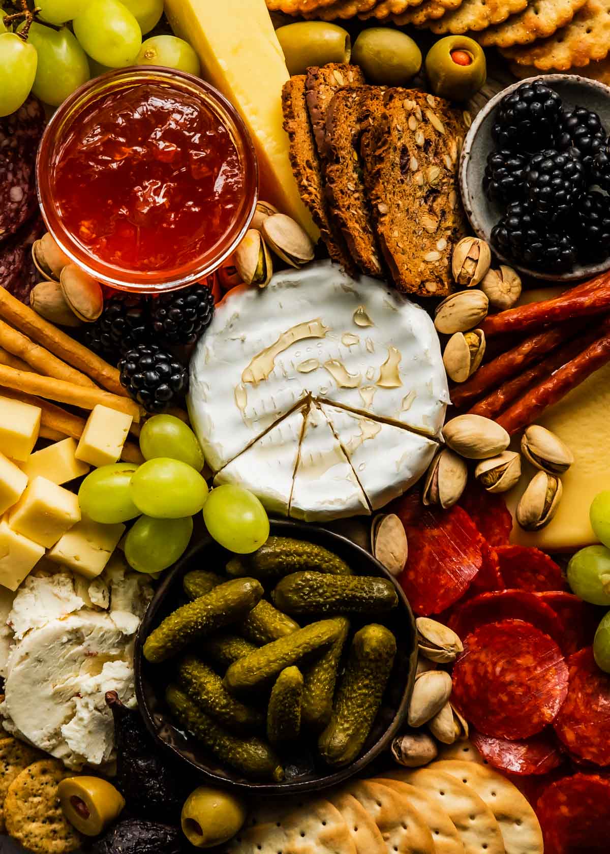 a closeup of a charcuterie board with brie drizzled with honey, cubed cheeses, crostini, jam, grapes, and nuts