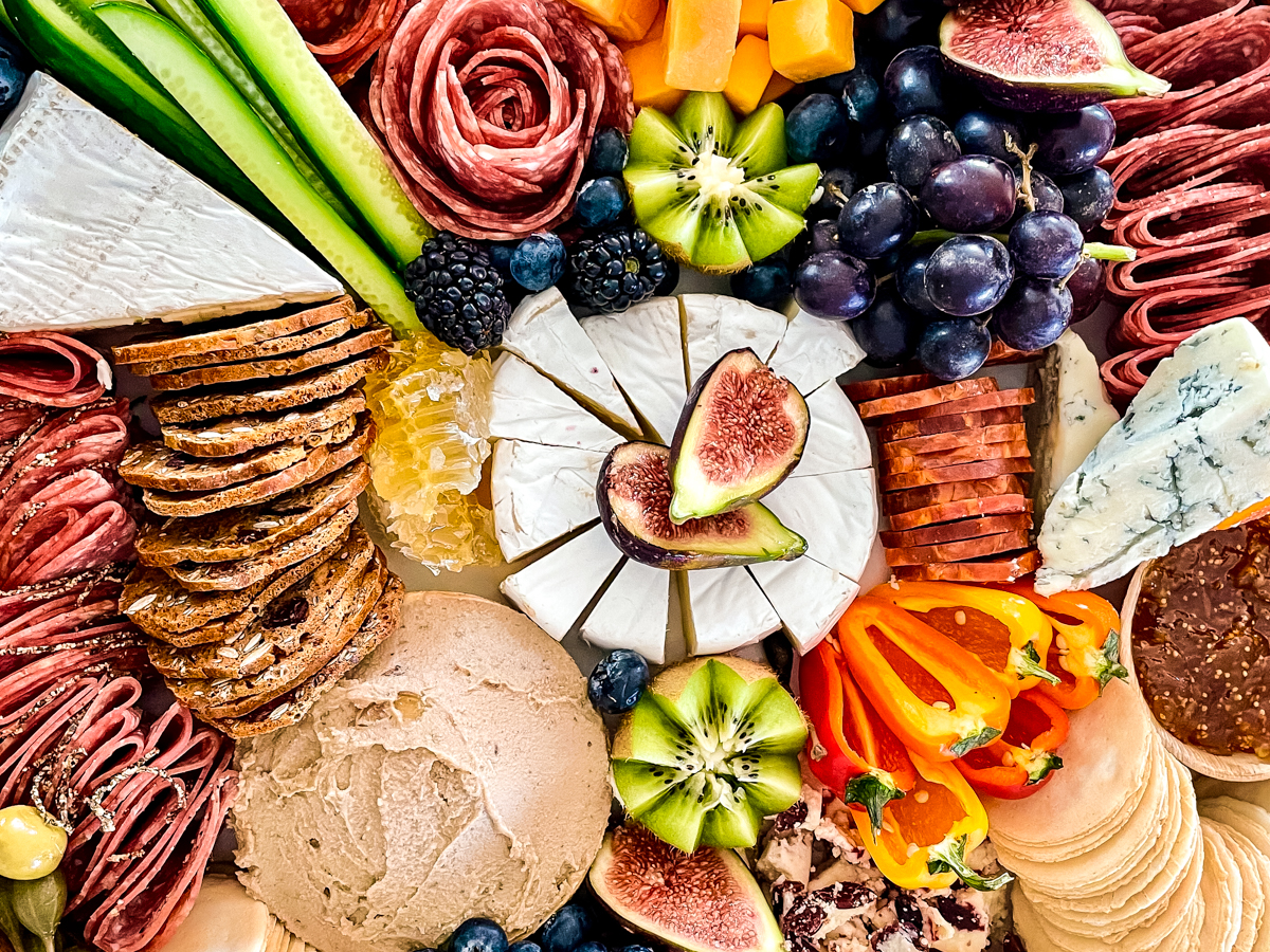 Top-down view of a salami charcuterie board with brie, fresh vegetables, duck liver pâté, and crackers