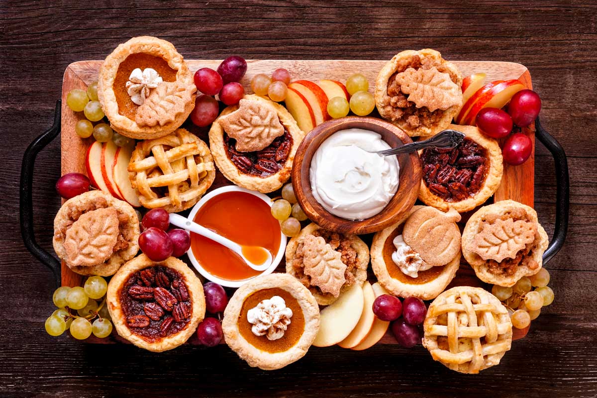 an overhead shot of a medium-sized, rectangle dessert charcuterie board with assorted mini-pies, apple slices, grapes, and two dipping sauces