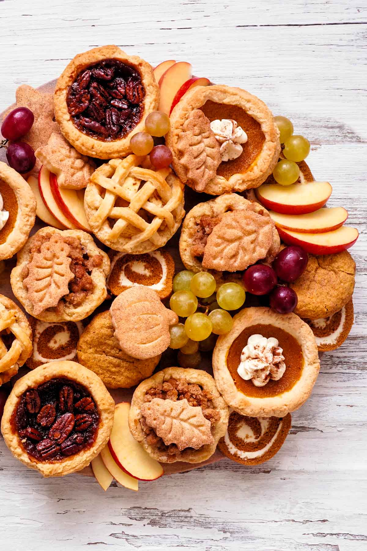 an overhead shot of a small, round dessert charcuterie board with assorted mini-pies, cookies, apple slices, and grapes