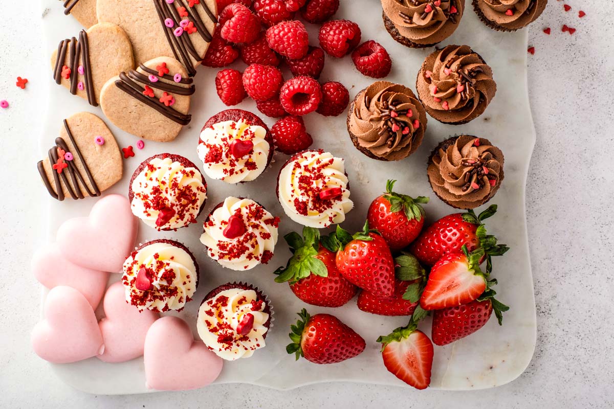 a small dessert charcuterie board with two kinds of mini-cupcakes (red velvet and chocolate) along with heart-shaped cookies and fresh raspberries and strawberries