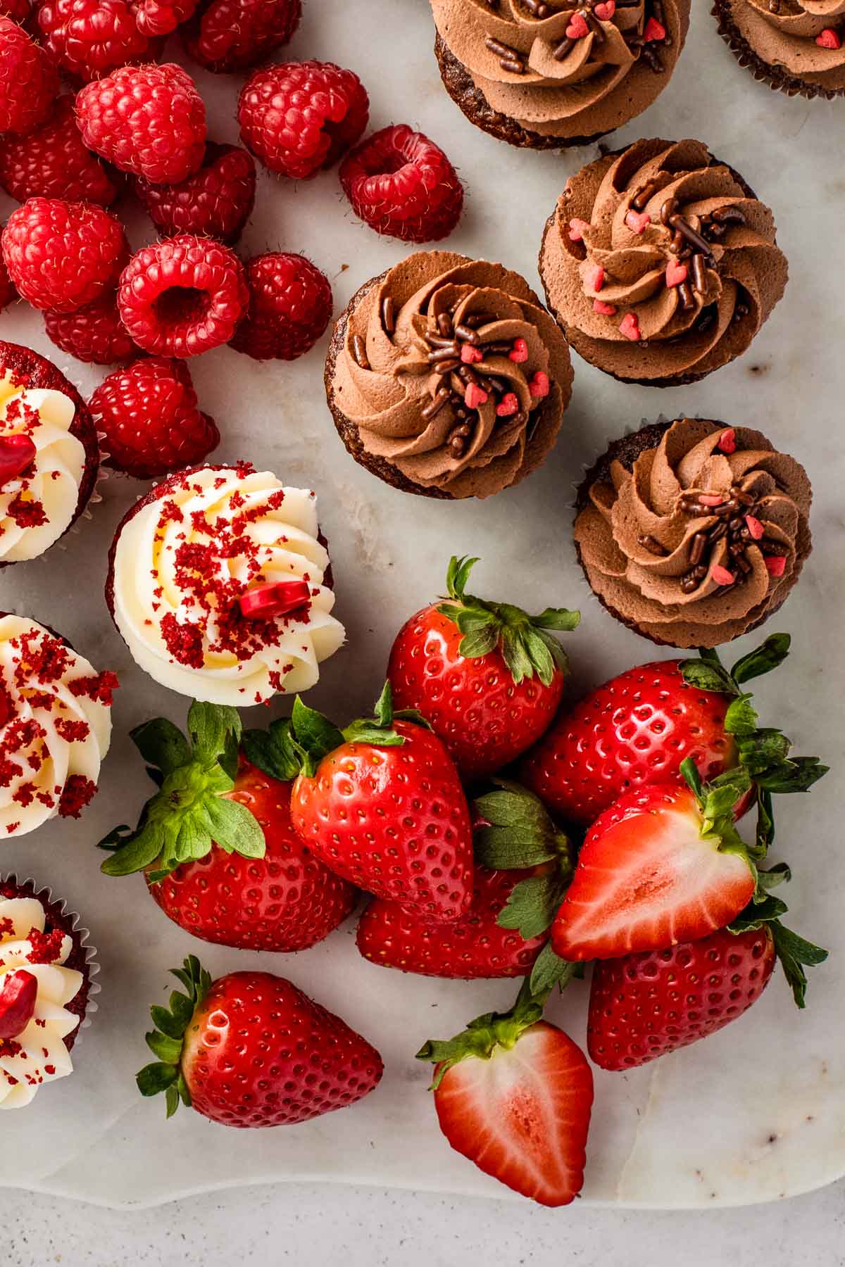 a closeup of a dessert charcuterie board with two kinds of mini-cupcakes (red velvet and chocolate) along with heart-shaped cookies and fresh raspberries and strawberries