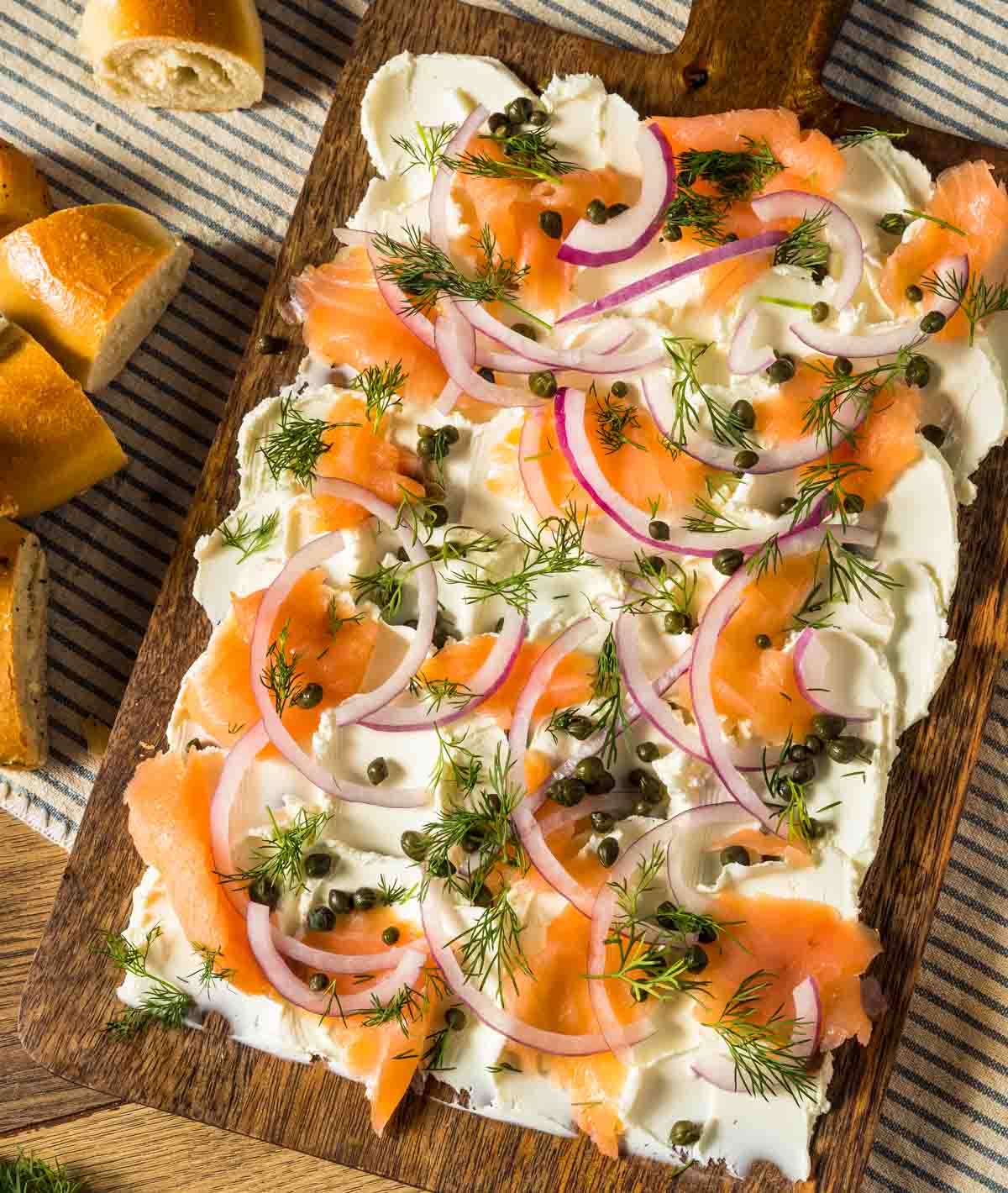 a bagel board with cream cheese, smoked salmon, red onion, capers and dill with cut up bagels around it