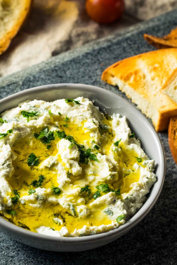 a gray bowl filled with boursin dip and topped with fresh herbs and olive oil, next to slices of toasted bread