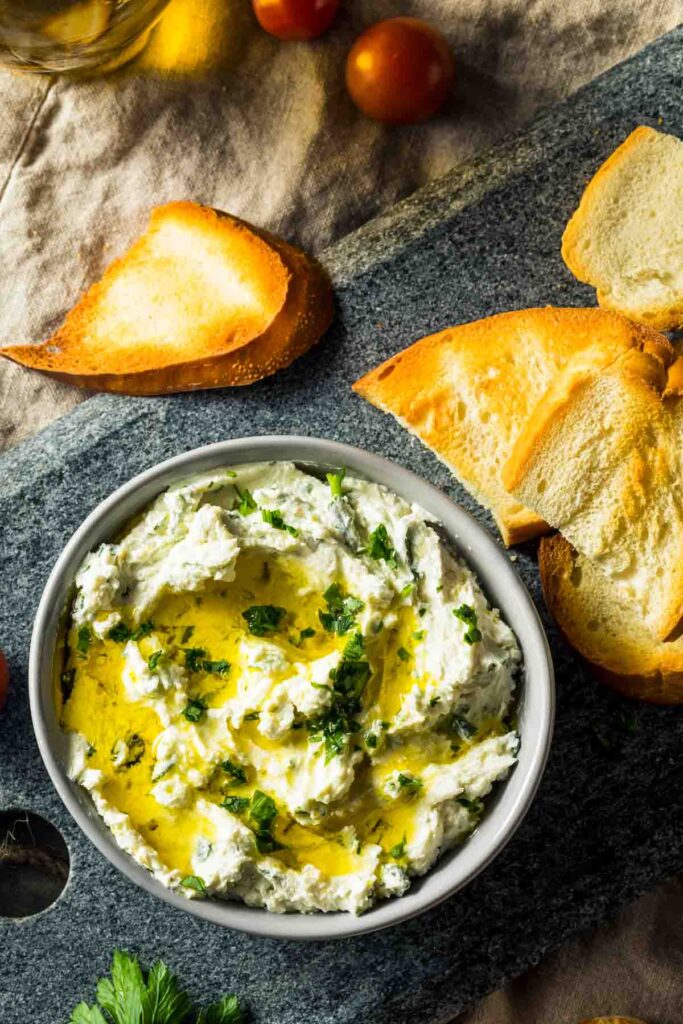 an overhead shot of a gray bowl on a cutting board filled with boursin dip and topped with fresh herbs and olive oil, next to slices of toasted bread