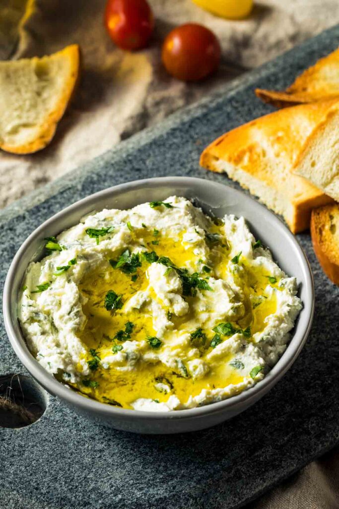 a gray bowl on a cutting board filled with boursin dip and topped with fresh herbs and olive oil, next to slices of toasted bread