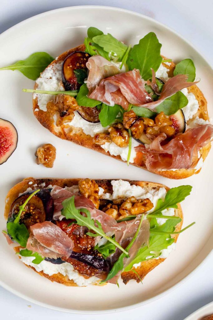 an overhead shot of  a white plate with two whipped ricotta crostini with figs, walnuts, and parma ham
