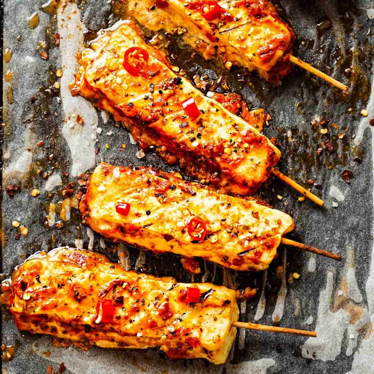 four skewers of hot honey halloumi cheese on parchment paper and garnished with thinly slice red chile peppers