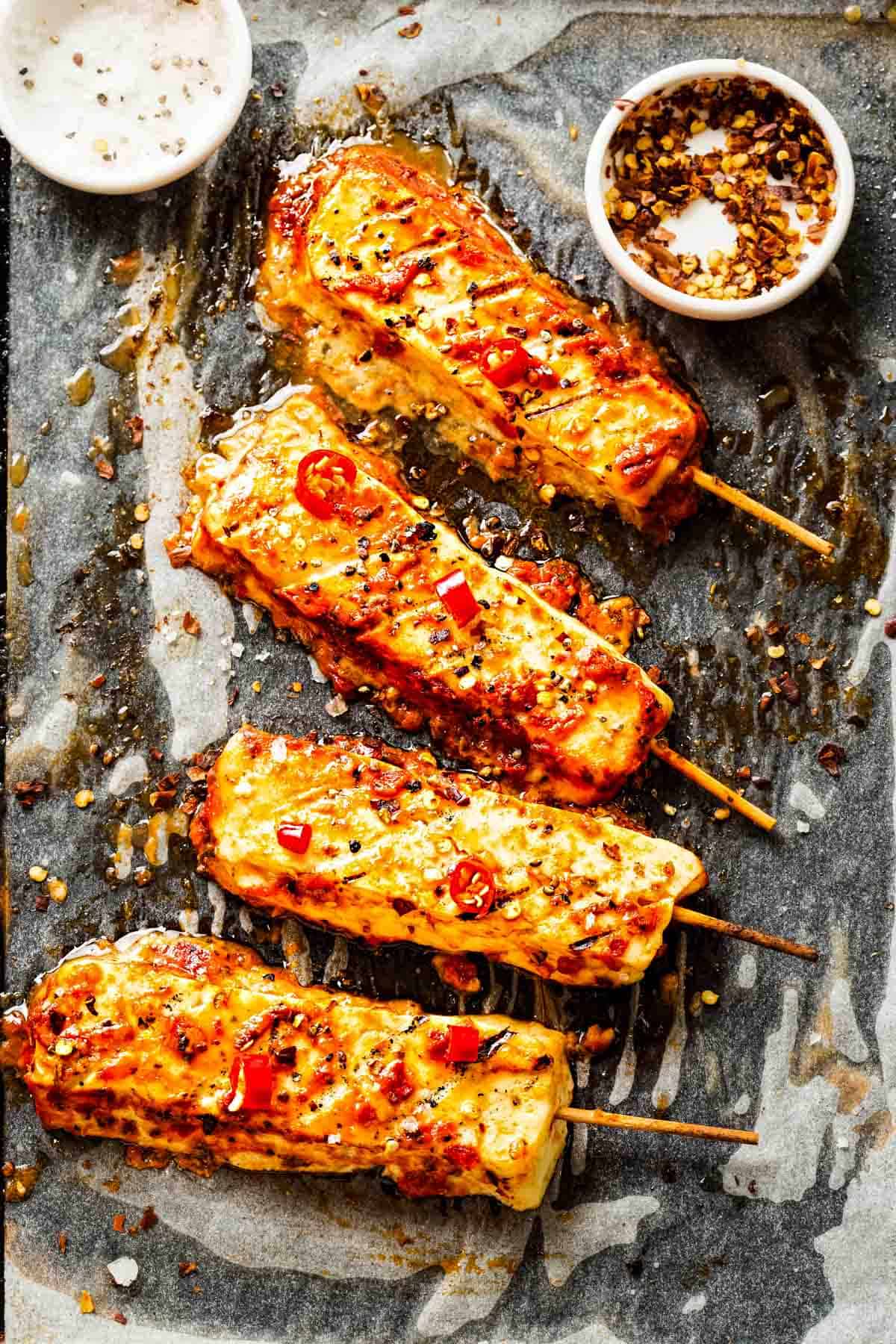 four skewers of hot honey halloumi cheese on parchment paper next to a tiny dish of crushed red pepper flakes