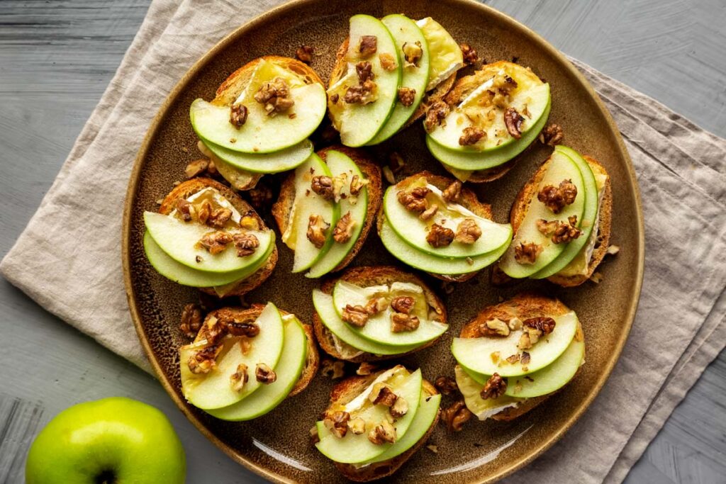 a large brown plate filled with apple and brie crostini with walnuts and drizzled honey on a folded tan linen napkin