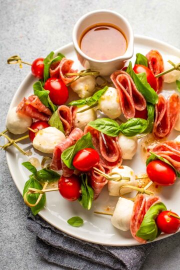 a large white plate filled with charcuterie skewers made with mozzarella, grape tomatoes, salami, and fresh basil with a dish of homemade italian dressing on the side