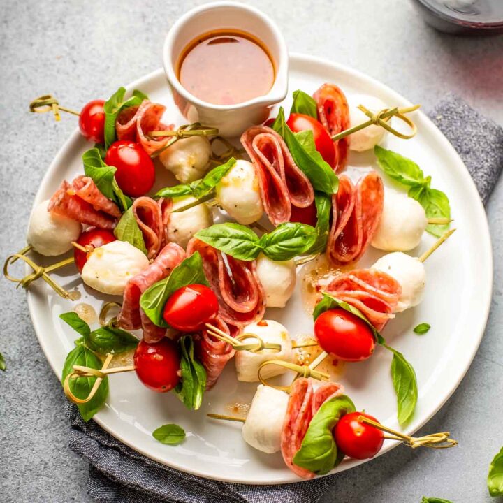 a large white plate filled with charcuterie skewers made with mozzarella, grape tomatoes, salami, and fresh basil with a dish of homemade italian dressing on the side next to a glass of red wine
