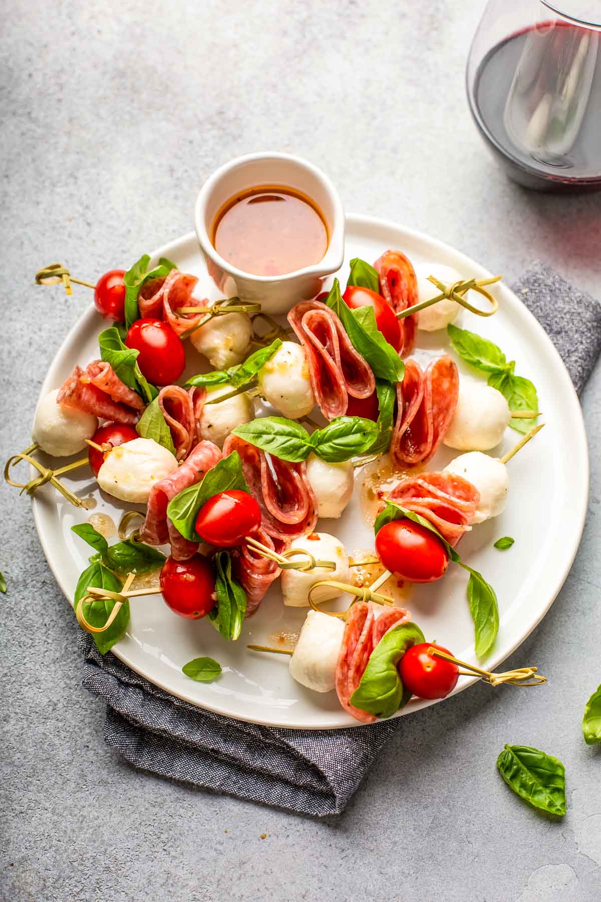 a large white plate filled with charcuterie skewers made with mozzarella, grape tomatoes, salami, and fresh basil with a dish of homemade italian dressing on the side next to a glass of red wine