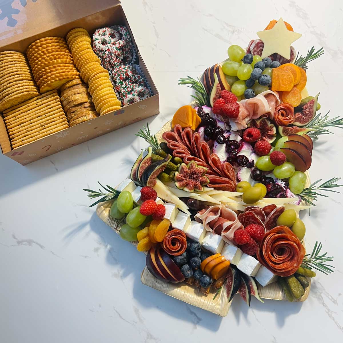 a christmas tree charcuterie board with brie, manchego, havarti, salami, prosciutto, cacciatore sausage, olives, fresh fruit, with pretzel sticks for the tree trunk and rosemary for the garnish next to a small box of crackers