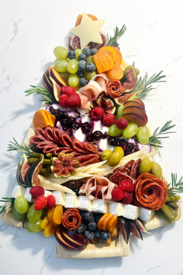 a christmas tree charcuterie board with brie, manchego, havarti, mild and spicy salami, prosciutto, cacciatore sausage, olives, fresh fruit, with pretzel sticks for the tree trunk and rosemary for the garnish