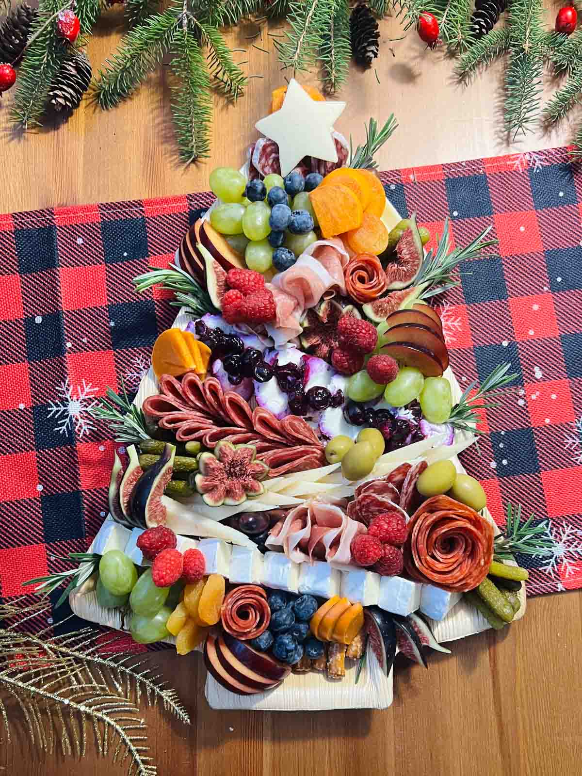 a christmas tree charcuterie board with brie, manchego, havarti, salami, prosciutto, cacciatore sausage, olives, fresh fruit, with pretzel sticks for the tree trunk and rosemary for the garnish on a red and green checkered napkin