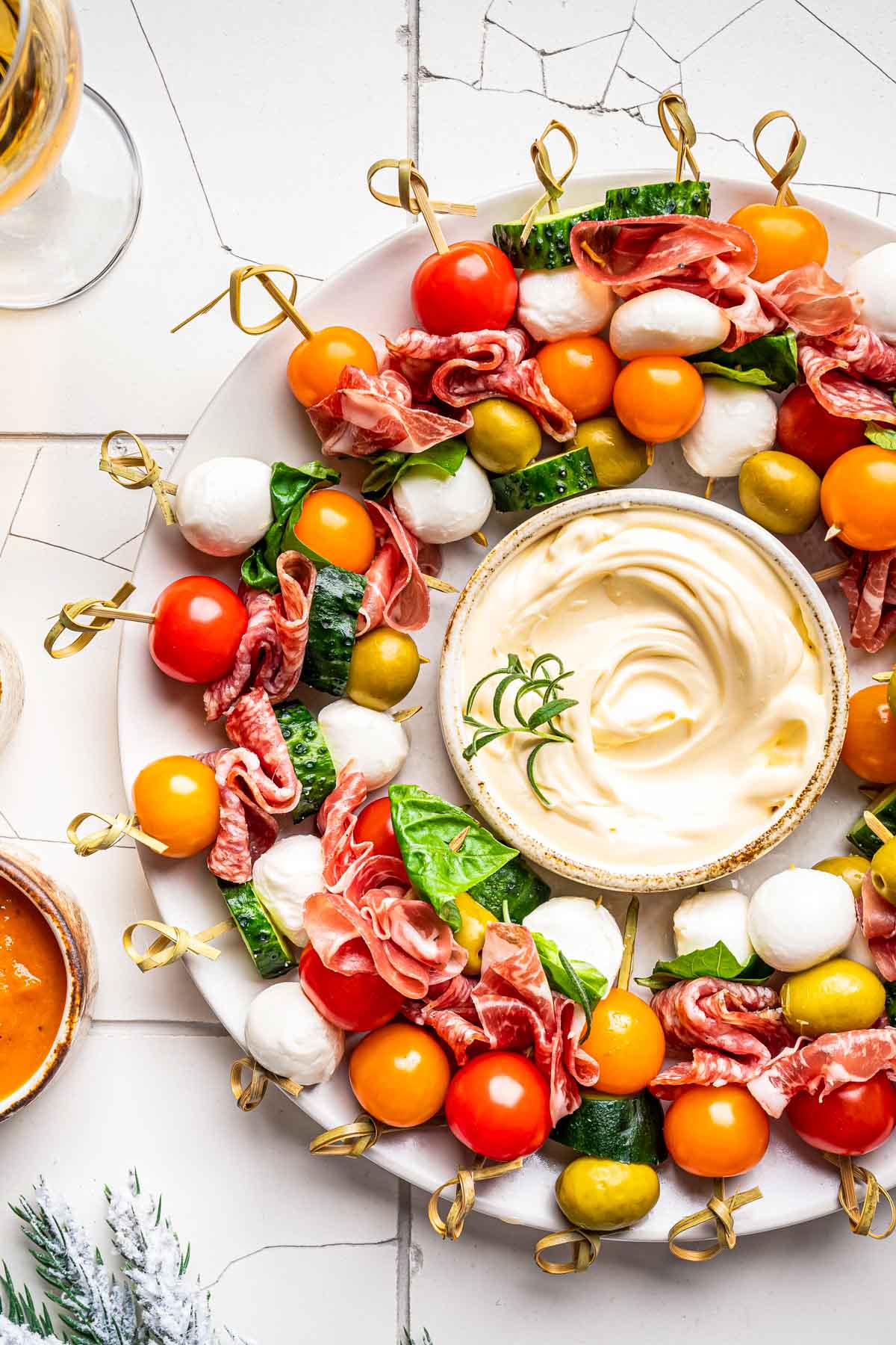 an overhead shot of a christmas wreath charcuterie board made up of small skewers of bocconcini, tomatoes, and prosciutto with a small bowl of dip in the center of a white plate