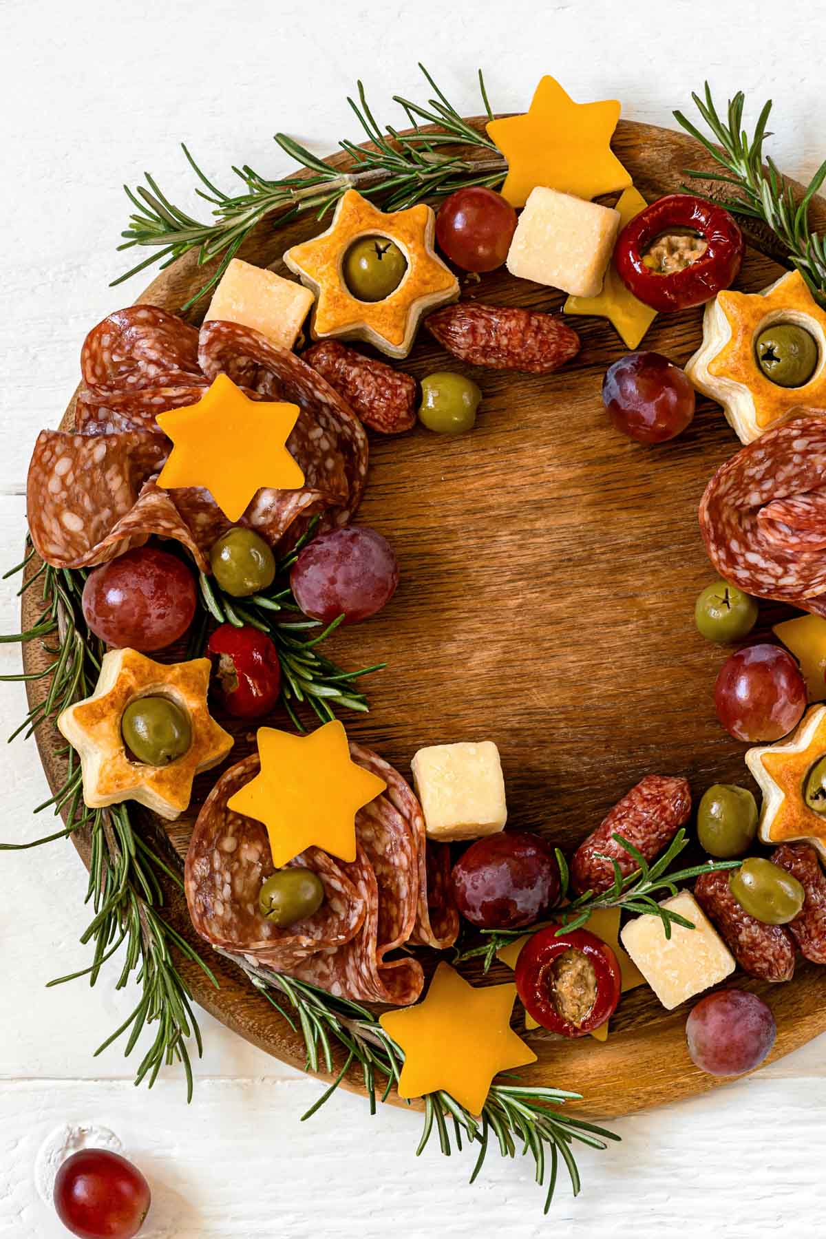 a christmas wreath charcuterie board made up of cured meats, orange cheese stars, star-shaped puff pastry, candied walnuts, red grapes and rosemary garnish