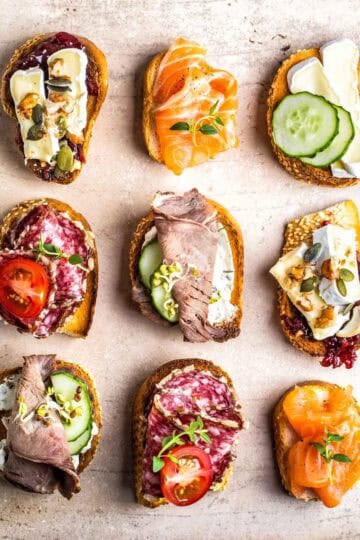 an assortment of nine crostini with different toppings: brie, pears, honey, blue cheese, salami, cucumbers, tomatoes, smoked salmon, and deli meat