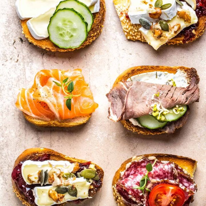 an assortment of six crostini with different toppings: brie, pears, honey, blue cheese, salami, cucumbers, tomatoes, smoked salmon, and deli meat