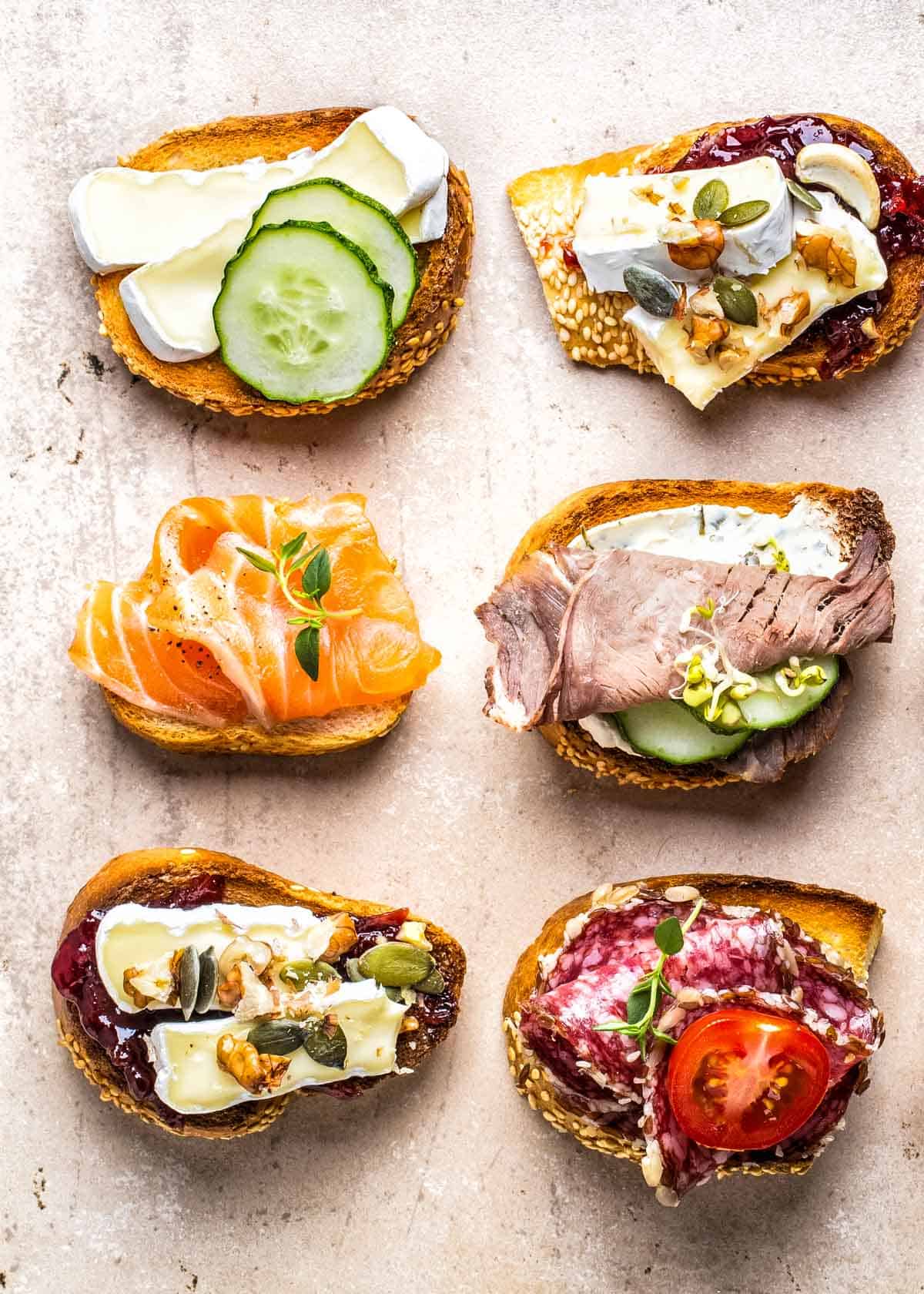 an assortment of six crostini with different toppings: brie, pears, honey, blue cheese, salami, cucumbers, tomatoes, smoked salmon, and deli meat