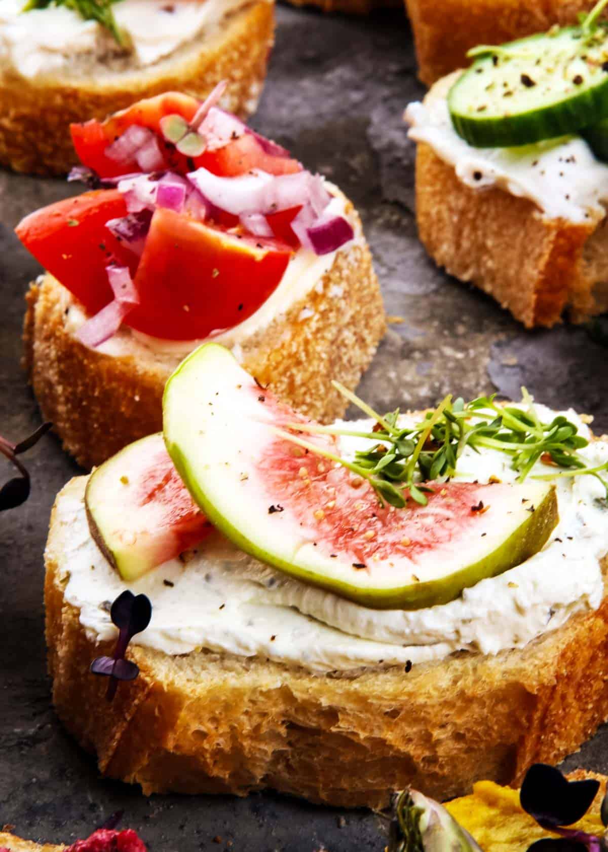a closeup of fig and ricotta crostini with assorted crostini in the background: tomatoes, whipped feta, cucumbers