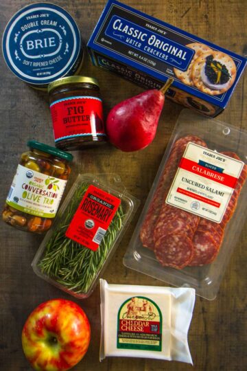all products needed for a $25 Trader Joe's charcuterie board: double cream brie, water crackers, fig jam, calabrese, olives, unexpected cheddar, an apple, a pear, and fresh rosemary